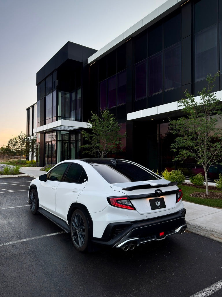A white high-performance sports sedan with black accents is parked in a lot in front of a modern black glass building at dusk.