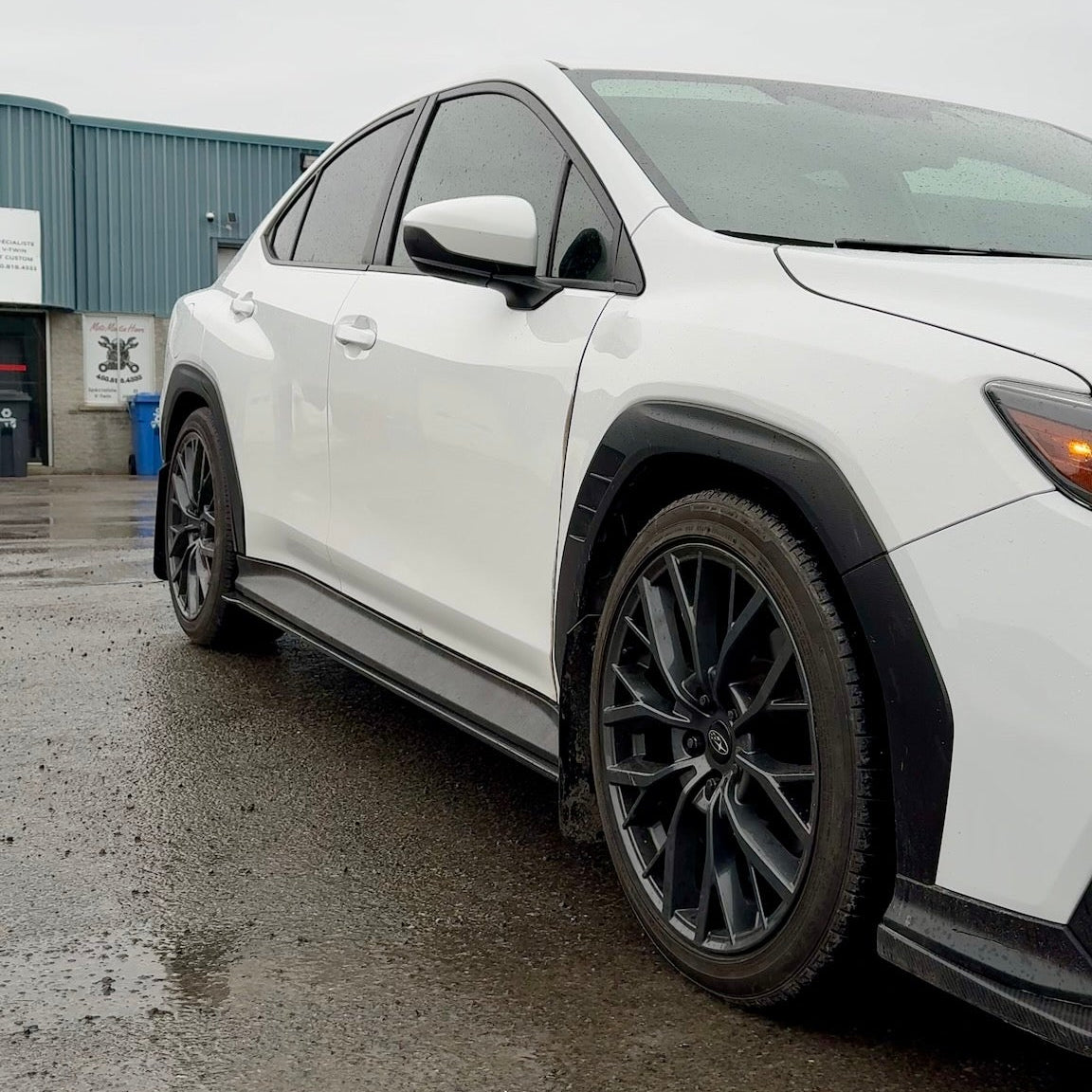 White car parked on a wet pavement with a building in the background