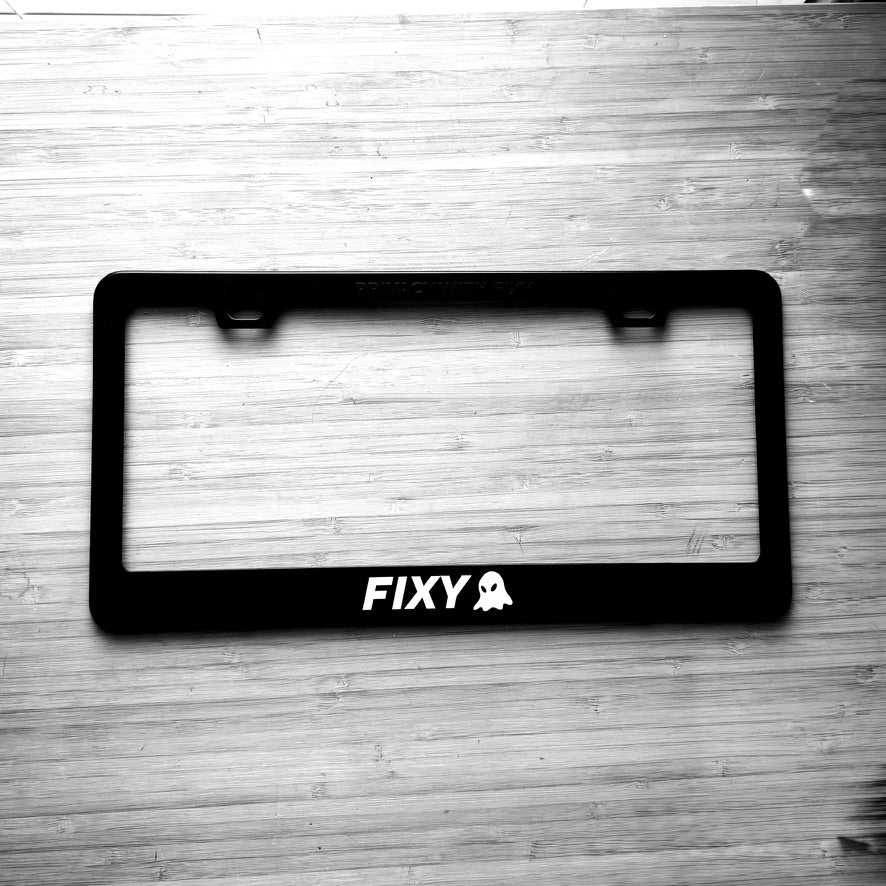 Black license plate frame with 'FIXY' branding on a wooden surface