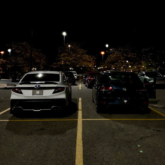 Dark parking lot with cars at night, illuminated by streetlights.