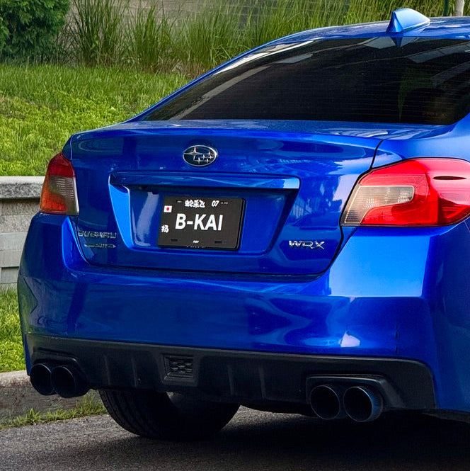 A blue Subaru WRX sedan photographed outdoors on a paved surface, showing the rear three-quarter view with grass, trees, and a brick wall in the background.