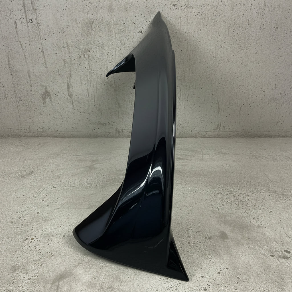 A glossy black car rear spoiler wing leaning against a light brick wall on a textured concrete floor.