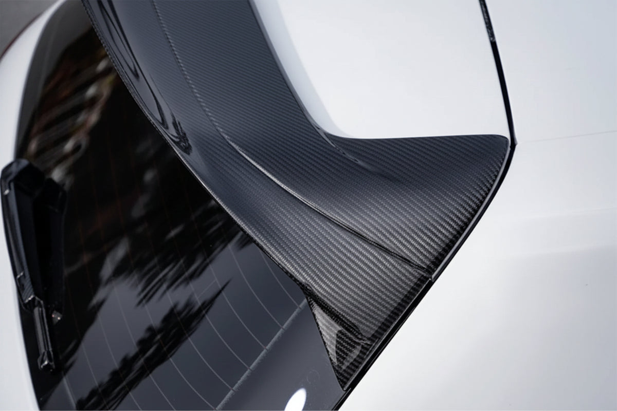 Close-up of a carbon fiber rear spoiler wing on a dark gray car, showing the textured weave pattern against a glossy body panel.