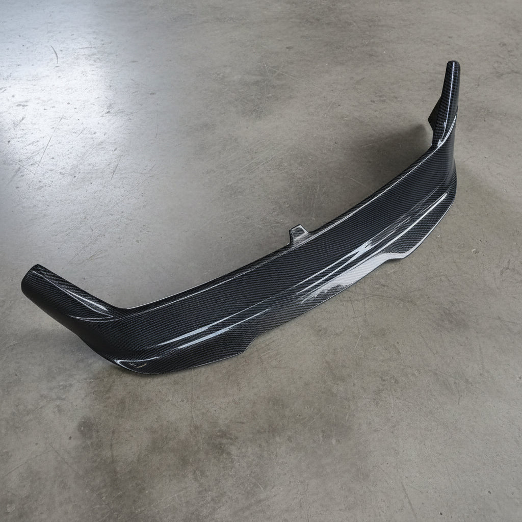 A glossy black carbon fiber automotive bumper lip or front splitter shown on a textured beige fabric background.