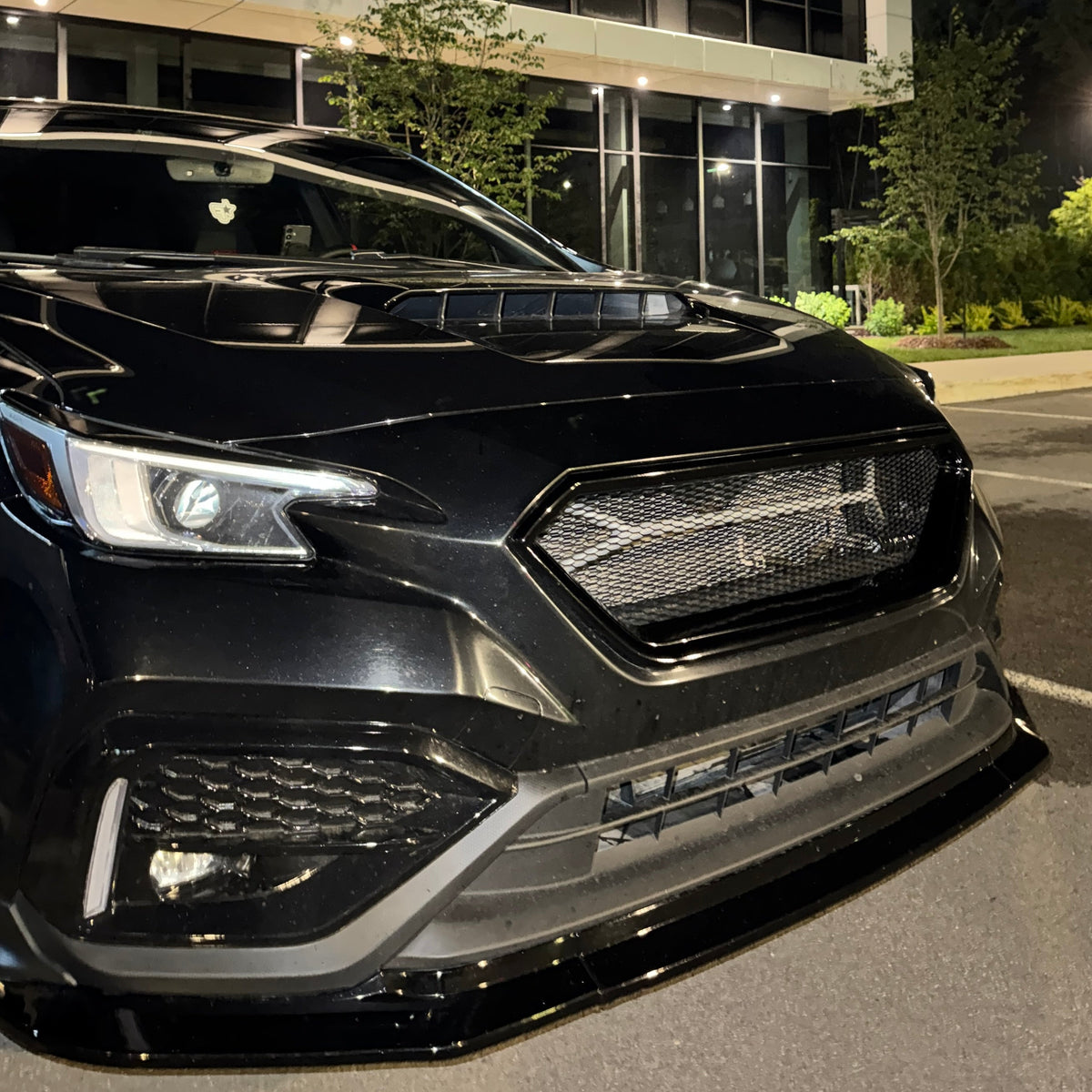 A black car with a custom front lip, mesh grille, and hood scoop, parked at night in front of a modern glass building.