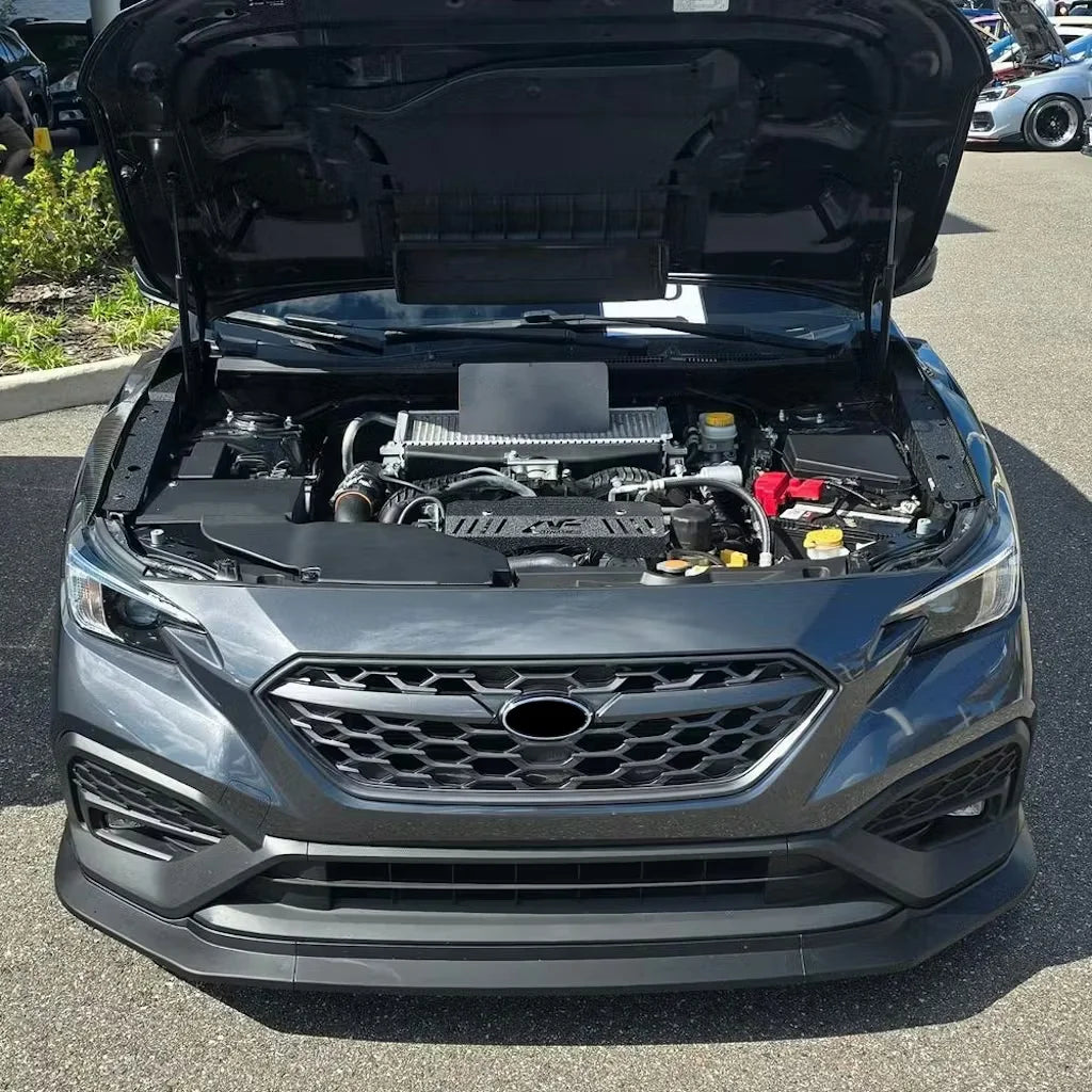 Front view of a dark gray Subaru SUV with the hood open, showing the engine bay, photographed outdoors on a paved surface.