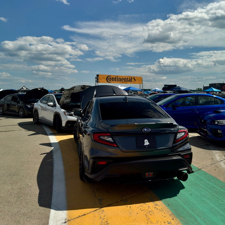 A black Subaru sedan photographed from the rear in an outdoor parking lot under a bright blue sky with scattered clouds. Other cars are visible in the background, along with a yellow Continental's sign.