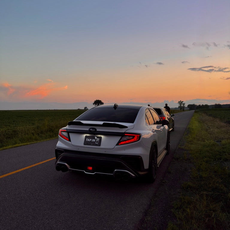 A white Subaru WRX sedan is shown from the rear on a rural road at sunset, with another vehicle behind it and open fields on both sides of the road.