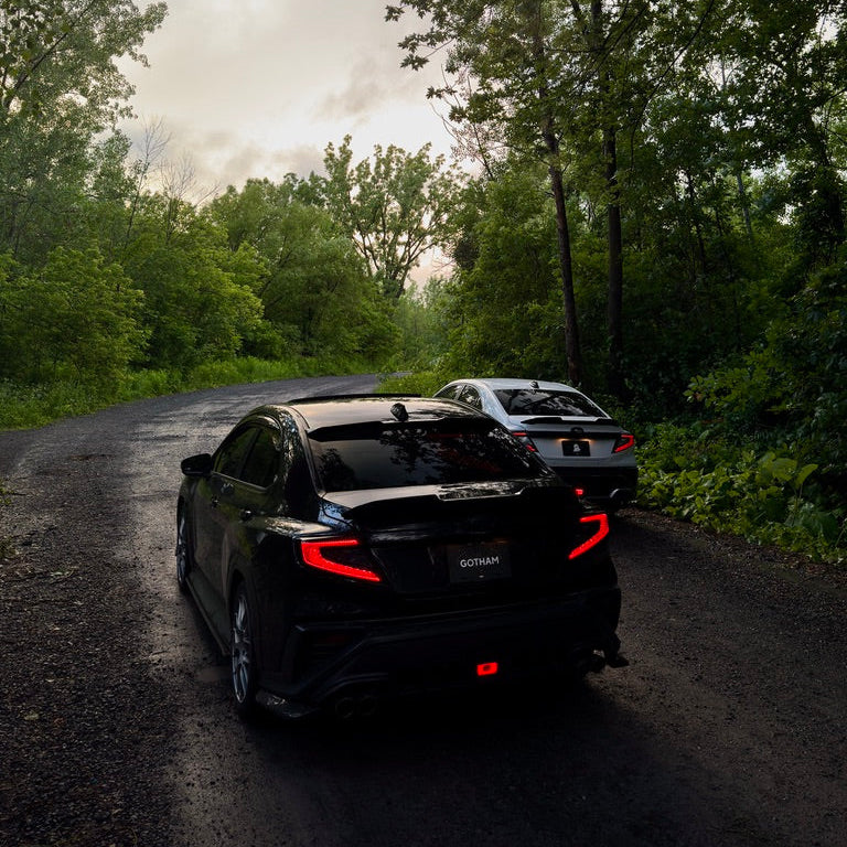 A black sedan photographed from the rear on a narrow forest road, with a second car behind it and dense green trees on both sides under an overcast sky.