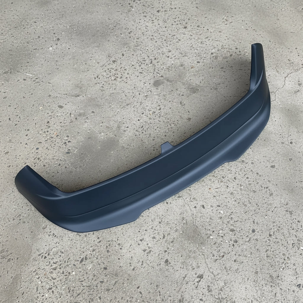A matte black plastic automotive rear bumper lip or spoiler trim piece, photographed from above on a textured beige carpet background.